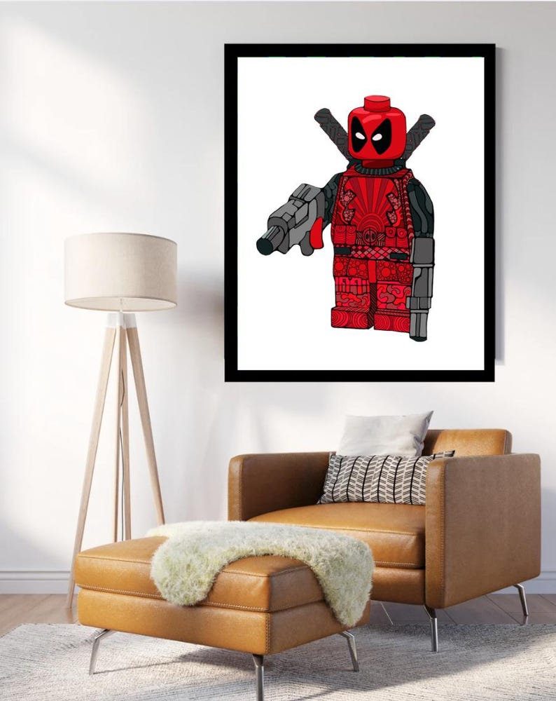 May include: A framed print of a red and black Lego minifigure of Deadpool. The minifigure is holding a gun and has a sword on its back. The background is white.