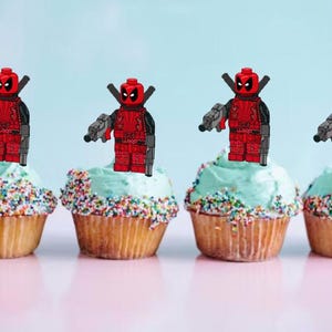 May include: Five cupcakes decorated with red and black Lego Deadpool figures holding guns. The cupcakes are topped with light blue frosting and colourful sprinkles.