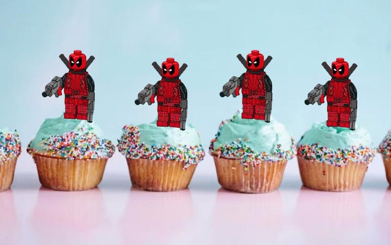 May include: Five cupcakes decorated with red and black Lego Deadpool figures holding guns. The cupcakes are topped with light blue frosting and colourful sprinkles.