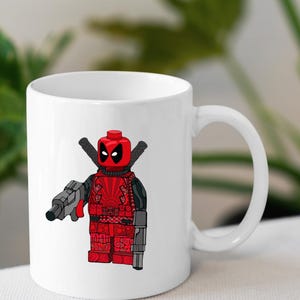 May include: White ceramic coffee mug featuring a red and black Lego Deadpool character holding a gun. The character is wearing a red and black suit with a black mask.