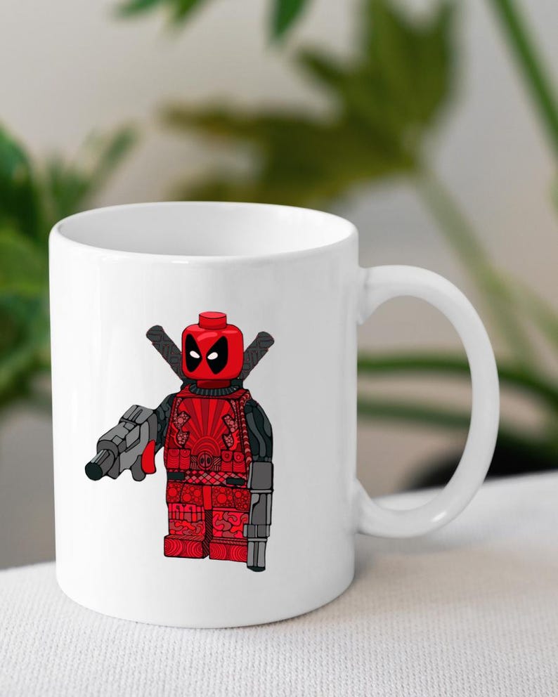 May include: White ceramic coffee mug featuring a red and black Lego Deadpool character holding a gun. The character is wearing a red and black suit with a black mask.