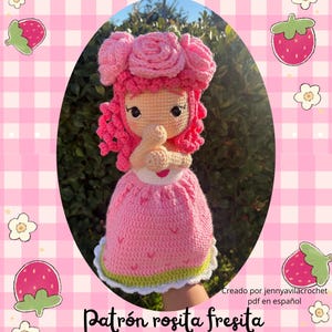 Puede incluir: Muñeca de ganchillo hecha a mano con un vestido de fresa rosa y un tocado a juego adornado con rosas. La muñeca tiene la piel clara, ojos negros y pelo rizado rosa. El fondo presenta un patrón de cuadros rosas con ilustraciones de fresas y flores.