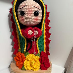 Puede incluir: Muñeca de ganchillo hecha a mano de Nuestra Señora de Guadalupe, con una combinación de colores rojo, verde, amarillo y negro. La muñeca tiene una expresión serena y está adornada con un rosario y flores de ganchillo. Está montada sobre una base de madera.