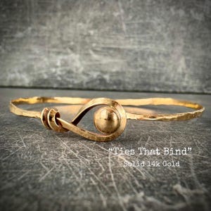 May include: A gold-colored bangle bracelet with a hammered texture. The bracelet features a unique design with a spiral and a sphere at the center. The text "Ties That Bind" and "Solid 14k Gold" are visible.