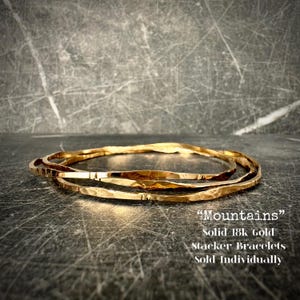 May include: Three gold-colored stacker bracelets with a textured, hammered finish. The bracelets are arranged in a layered style, with the text "Mountains" and "Solid 18k Gold" visible in the image. The bracelets are sold individually.