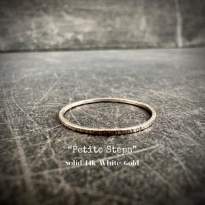 May include: A delicate, textured ring made of solid 14k white gold. The ring is thin and circular, with the words "Petite Steps" and "Solid 14k White Gold" in white text. The ring is set against a dark, textured background.