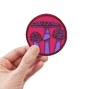 May include: A pink and purple embroidered patch featuring three mushrooms. The mushrooms have a dark brown cap and a purple stem.