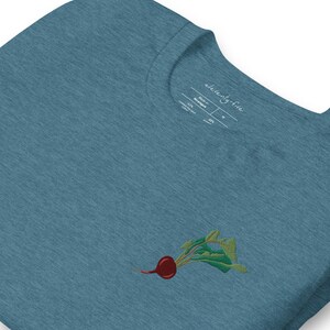 May include: A teal blue t-shirt with an embroidered red beet with green leaves. The t-shirt has a white tag that says "eleventy five" and "Made in Nicaragua".