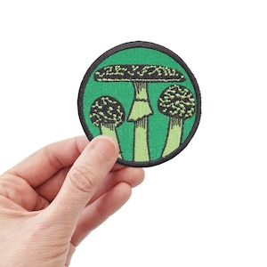 Puede incluir: Un parche bordado verde con un diseño de hongo negro y blanco. El hongo tiene un sombrero grande y un tallo grueso. Hay dos hongos más pequeños a cada lado del hongo principal.