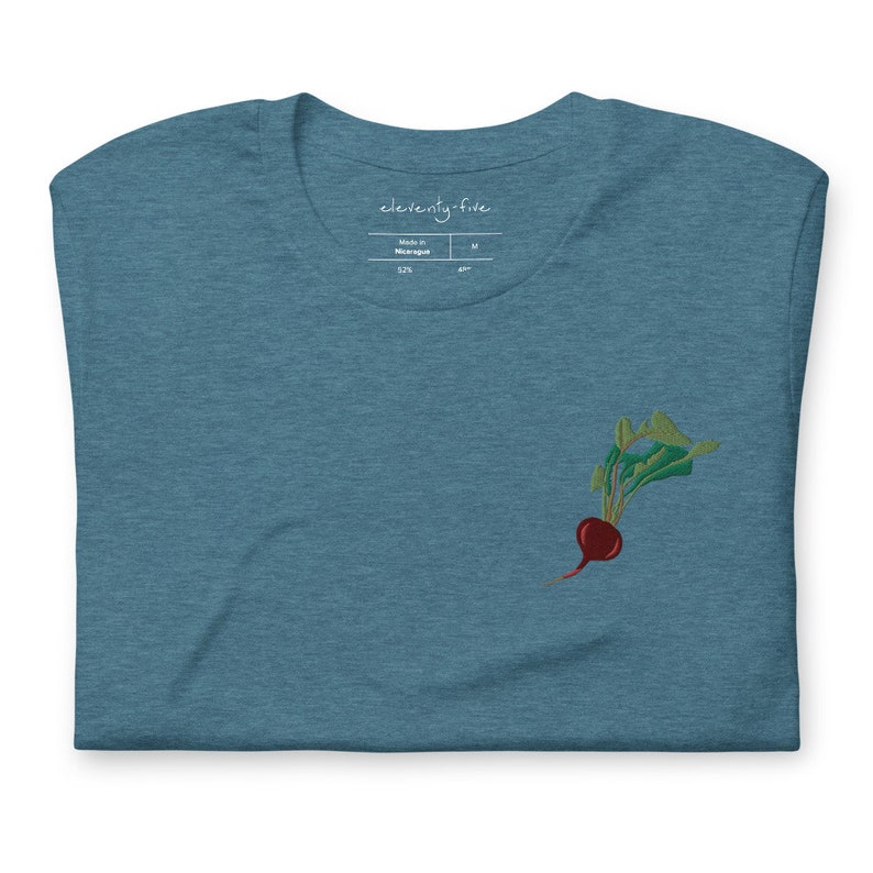 May include: A teal blue t-shirt with an embroidered beet design. The shirt has a tag that says "eleventy-five" and "Made in Nicaragua".