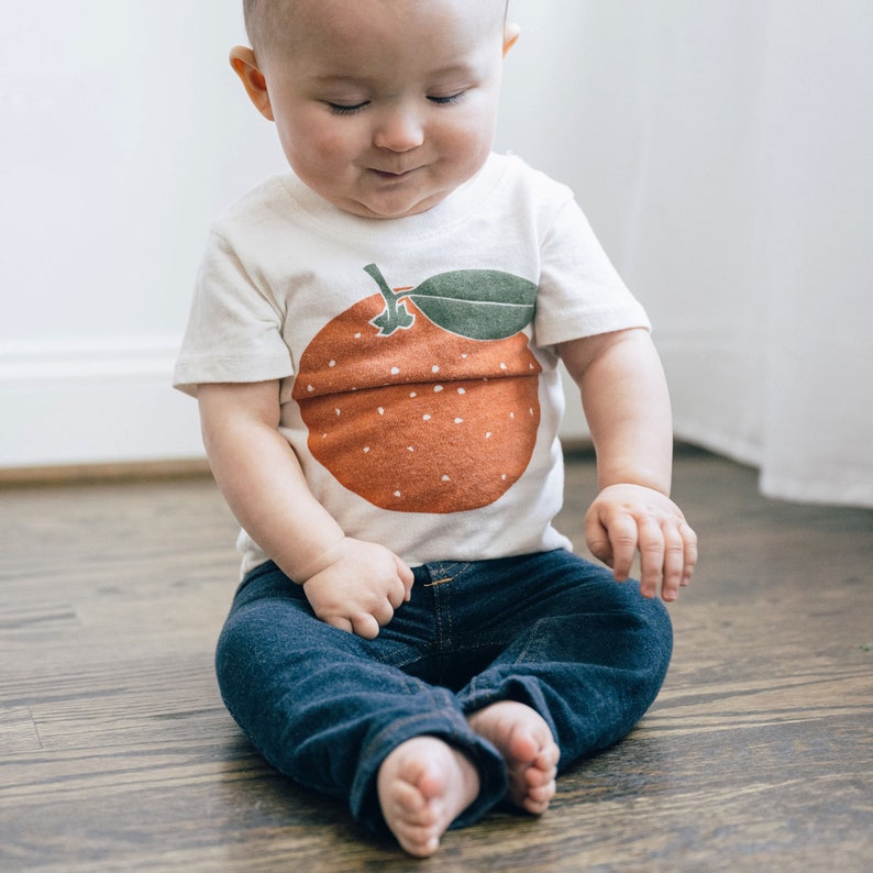 May include: A toddler wearing a white t-shirt with an orange fruit graphic and blue jeans. The fruit has a green leaf.