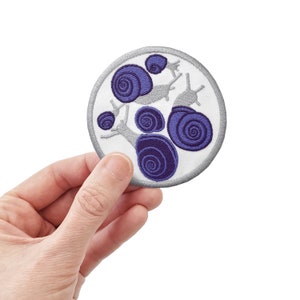 May include: A white circular patch with an embroidered design of purple snails. The snails are arranged in a cluster with their shells facing outward.