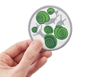 May include: A white and gray circular embroidered patch with a design of green snails. The snails are arranged in a cluster, with their shells facing outward.