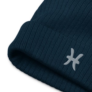 Könnte beinhalten: Eine dunkelblaue Strickmütze mit einem weißen gestickten Sternzeichen Fische.