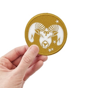 May include: A gold embroidered patch with a white ram's head and the astrological symbol for Aries. The patch has a white outline and three white stars.