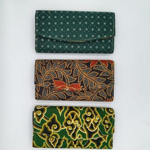Può includere: Tre portafogli in tessuto con chiusure a patta. Il portafoglio superiore è verde scuro con pois bianchi. Il portafoglio centrale ha uno sfondo nero con motivi a foglie arancioni e marroni. Il portafoglio inferiore è verde con disegni a forma di nuvola gialla e nera.