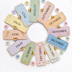 May include: A circular arrangement of twelve rectangular month labels, each with a gold-colored clip, featuring pastel colors and unique patterns. Twelve small circular number tokens from 1 to 12 are interspersed between the month tags.