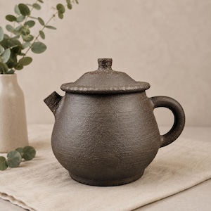 Op de afbeelding: Een donkerbruine keramische theepot met een textuuroppervlak, een deksel en een gebogen handvat. De theepot staat op een beige doek, met een vaas en groen in de achtergrond.
