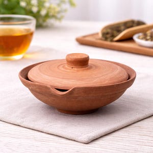 May include: A terracotta tea bowl with a matching lid. The bowl has a rounded shape with a small spout and sits on a light-coloured cloth. The lid has a small knob for easy handling. A glass of tea is visible in the background.
