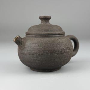 Puede incluir: Pequeña tetera de cerámica marrón oscuro con tapa y asa curva. La tetera tiene una superficie texturizada y un pico. La tapa tiene un pequeño pomo en la parte superior. Diseñada para preparar té.