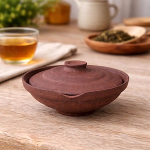 Puede incluir: Un pequeño cuenco de té de cerámica de color marrón rojizo con una tapa a juego. El cuenco tiene una forma redondeada con un borde ligeramente ensanchado y un pequeño pico. La tapa tiene un pomo redondeado para facilitar su manejo. El cuenco está sobre una superficie de madera.