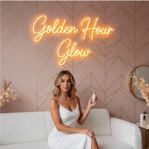 Puede incluir: Una mujer con un vestido blanco sentada en un sofá blanco sostiene un producto para el cuidado de la piel. El fondo presenta una pared rosa con un patrón geométrico y un letrero de neón que dice "Golden Hour Glow."