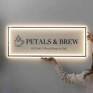 May include: Illuminated rectangular sign with the text "PETALS & BREW" in black, featuring a coffee cup and flower logo. The sign also includes "EST. 2025 | Floral Design & Café". The sign is being held up against a light gray wall.