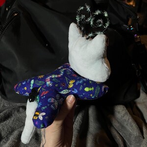 May include: A white plush toy with a blue and green fabric star-shaped body. The star is covered in a pattern of insects, flowers, and leaves. The toy is wearing a sparkly black crown.