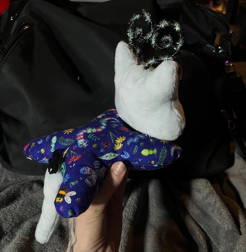 May include: A white plush toy with a blue and green fabric star-shaped body. The star is covered in a pattern of insects, flowers, and leaves. The toy is wearing a sparkly black crown.