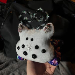 May include: A white plush toy with black eyes and a sparkly silver center. The toy has a blue and purple fabric on the back with a floral pattern. The toy has a sparkly silver and black wire antennae on top.