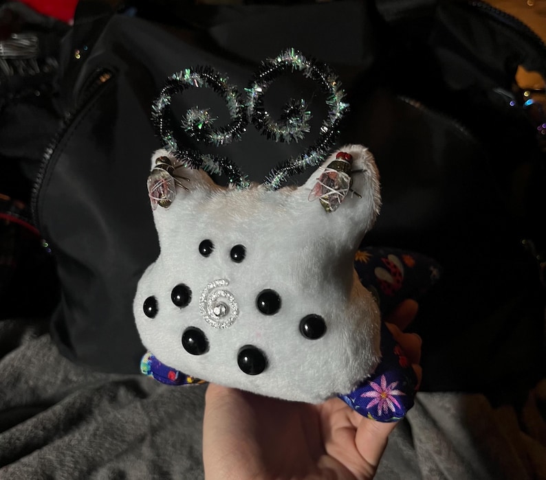 May include: A white plush toy with black eyes and a sparkly silver center. The toy has a blue and purple fabric on the back with a floral pattern. The toy has a sparkly silver and black wire antennae on top.
