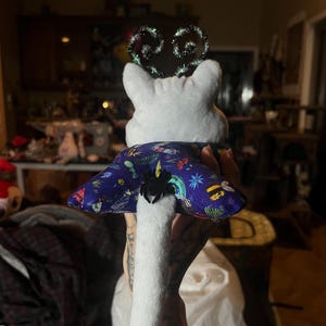 May include: White plush toy with a long tail, wearing a blue and purple fabric cape with a spider and butterfly design. The toy has a green and black sparkly headband.
