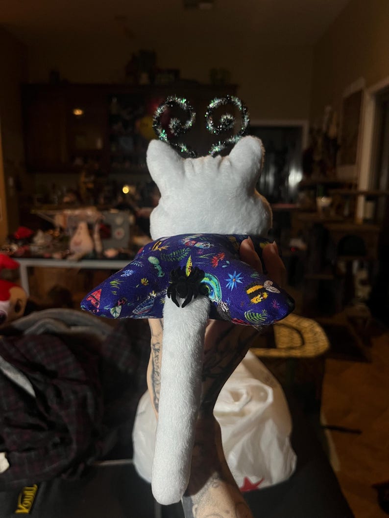 May include: White plush toy with a long tail, wearing a blue and purple fabric cape with a spider and butterfly design. The toy has a green and black sparkly headband.
