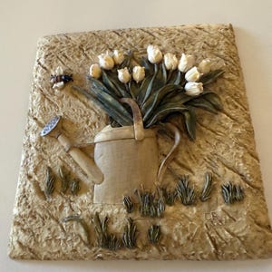 May include: A beige relief sculpture featuring a watering can overflowing with white tulips. A small bee and gardening tool are also depicted. The background has a textured, earthy appearance, with blades of grass. The artist's initials "CB" are visible.