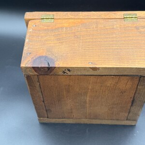 May include: A small, handcrafted wooden box with a hinged lid. The box is made of light brown wood and features brass hinges. The wood grain is visible, and the box appears to be well-constructed. The box is likely used for storage.