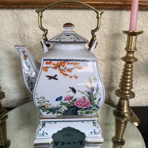 Peut inclure: Une théière en porcelaine blanche avec une poignée et des accents dorés. La théière présente un motif floral et d'oiseaux avec des teintes roses, oranges et bleues. La théière repose sur un support assorti avec une bordure dorée.