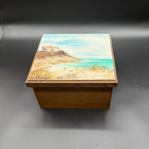 May include: A wooden box with a painted landscape scene on the lid. The artwork depicts a small house on a sandy shore with turquoise water and a blue sky. The box is rectangular and made of a dark wood.