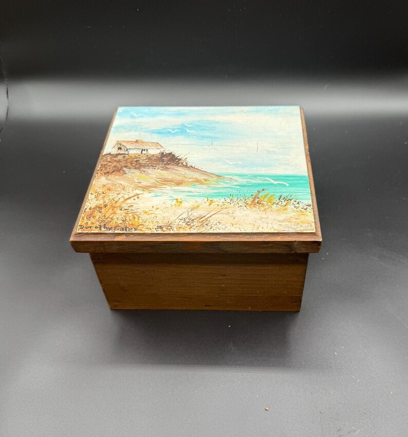 May include: A wooden box with a painted landscape scene on the lid. The artwork depicts a small house on a sandy shore with turquoise water and a blue sky. The box is rectangular and made of a dark wood.