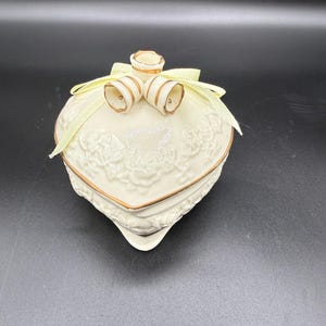 May include: Cream-colored heart-shaped trinket box with gold trim and a floral relief design. The lid features two bell-shaped ornaments and a pale yellow ribbon. A decorative piece for storing small items.