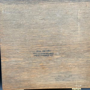 May include: A rectangular wooden box with a hinged lid. The wood has a natural grain pattern and a warm, brown tone. Text is printed on the wood. The box appears to be vintage or antique.