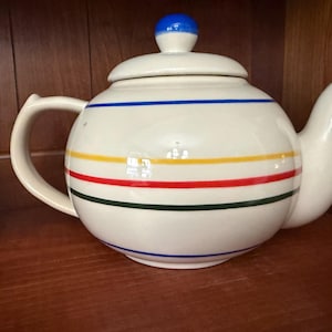 May include: A white ceramic teapot with a rounded body and a curved spout. The teapot features horizontal stripes in blue, yellow, red, and green. The lid has a blue knob. The teapot is displayed on a wooden surface.