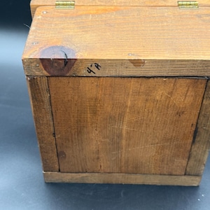 May include: A small, rectangular wooden box with a hinged lid. The box is constructed from light brown wood and features visible wood grain. The lid is attached with brass hinges. The text "4-A" is written on the side of the box.
