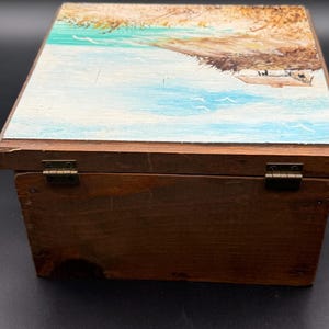 May include: A wooden box with a hinged lid. The lid is painted with a coastal scene featuring blue water, a sandy shore, and a small structure. The box is stained a dark brown color and has brass hinges.