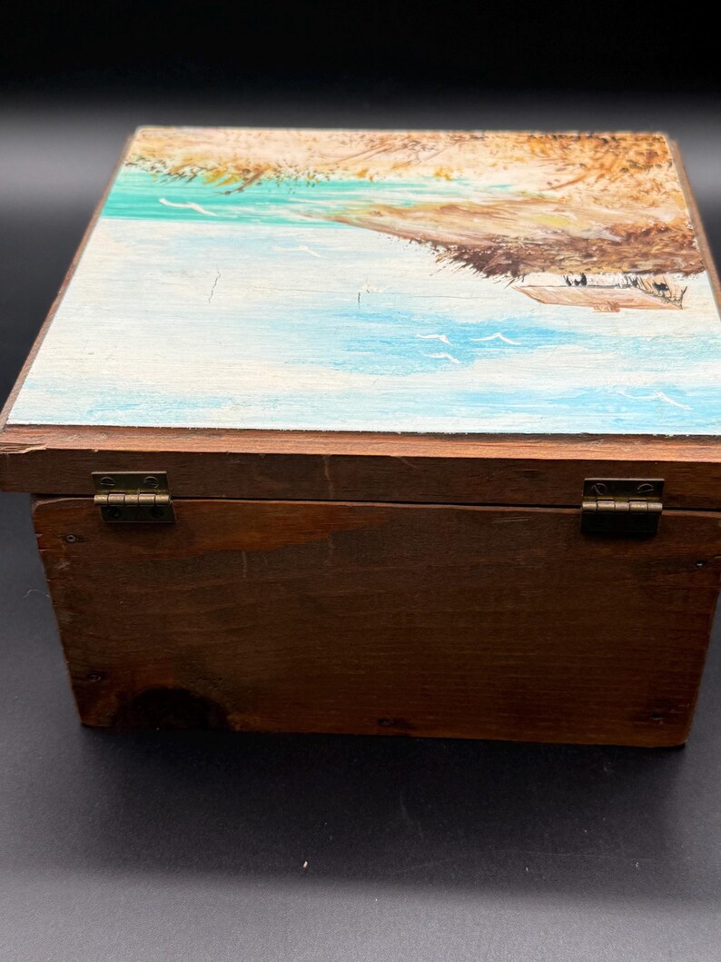May include: A wooden box with a hinged lid. The lid is painted with a coastal scene featuring blue water, a sandy shore, and a small structure. The box is stained a dark brown color and has brass hinges.