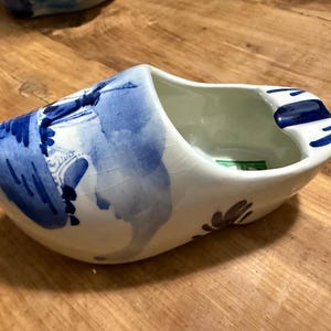 May include: A white ceramic Dutch shoe with blue and white painted designs. The shoe is shaped like a traditional clog and has a wide opening at the top.
