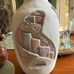 Può includere: Grande vaso in ceramica bianco sporco con un rilievo scolpito di un villaggio. Il rilievo presenta edifici multicolori e dettagli strutturati. Il vaso ha un corpo largo e un collo stretto, adatto per l'arredamento della casa.