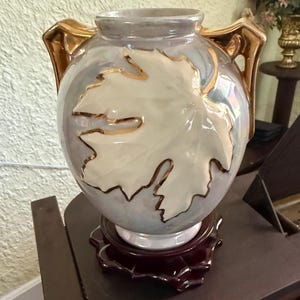 May include: A pearlescent ceramic vase with a large leaf design. The vase has gold-colored handles and leaf outlines. It sits on a dark red wooden stand. The vase is approximately 10 inches tall.