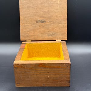 May include: Open wooden box with a hinged lid. The box is made of light brown wood with a yellow interior. The lid is open, revealing the inside of the box. The box appears to be empty.