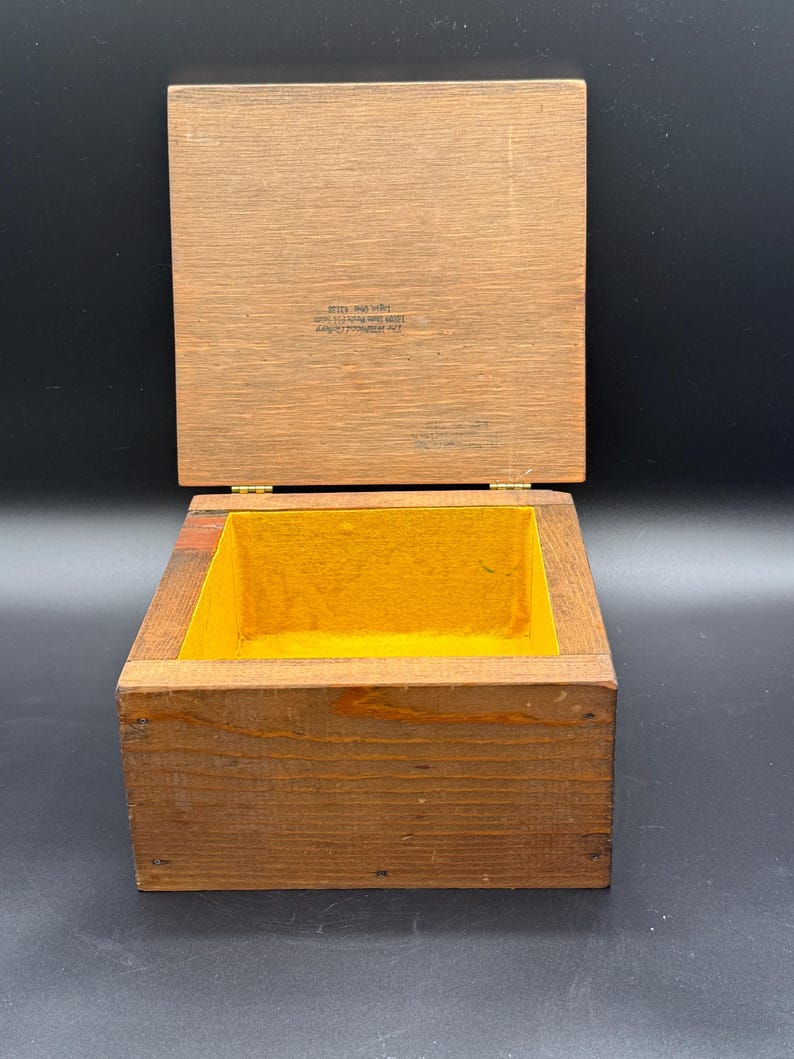 May include: Open wooden box with a hinged lid. The box is made of light brown wood with a yellow interior. The lid is open, revealing the inside of the box. The box appears to be empty.