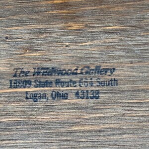 May include: Close-up of a wooden surface with the text "The Wildwood Gallery" and address details: 18809 State Route 664 South, Logan, Ohio 43138. The wood grain is visible.